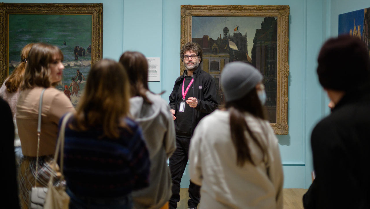 A museum assistant wearing a lanyard stands in front of a painting talking to an audience of young people standing in the gallery