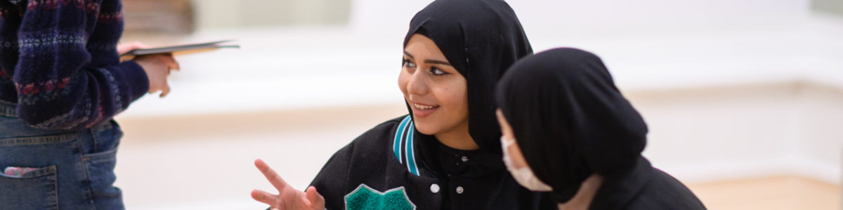 Two young Muslim women sitting and talking in a museum gallery with paintings on the wall.