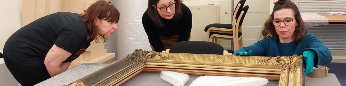 Three women at a Museum Basics training looking at the back of a framed picture on a table to prepare for packing it into tissue paper.