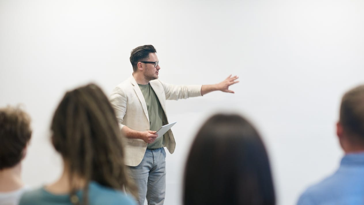 A man stands in front of a white wall talking and gesturing. Four people sit in front of him with their backs to the camera