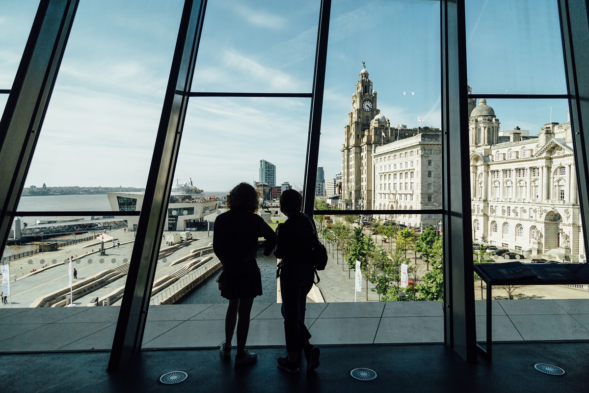 Two people in front of a large glass window overlooking Liverpool waterfront